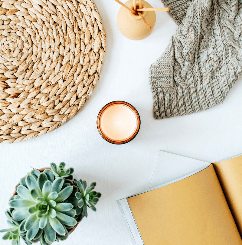 Flat lay image of a candle, succulent, wicker place mat, knit blanket and opened book.